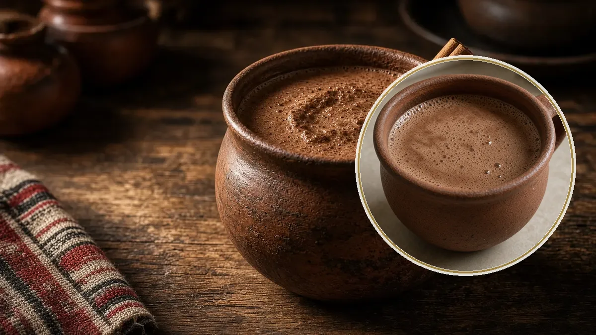 Una taza de barro contiene atole de chocolate espeso y espumoso, con una raja de canela en el borde, sobre madera oscura.